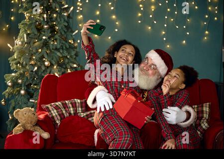 der weihnachtsmann und zwei afroamerikanische Schwestern machen zu Hause Selfie auf der roten Couch Stockfoto