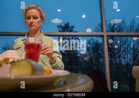 Eine Frau mit lockigem Haar sitzt an einem Café-Tisch und genießt ein buntes Getränk, mit einem Teller mit Essen vor dem Tisch, was ein warmes und einladendes Ambiente schafft Stockfoto