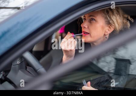 Eine junge Frau mit lockigen Haaren trägt Lippenstift in den Autospiegel auf, reflektiert ihre Schönheitsroutine während der Fahrt und hebt die Mischung aus Selbstpflege und d hervor Stockfoto