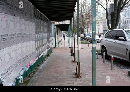 Eine Person schlendert einen ruhigen Bürgersteig entlang, neben Wänden, die mit Plakaten bedeckt sind. Kiew, Ukraine. November 2025. Stockfoto