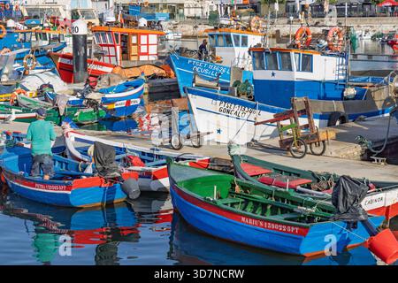 Geschäftige Fischerhafenszene in Setubal, Portugal, mit farbenfrohen Booten. Ein Fischer arbeitet in seinem kleinen blauen Boot am Pier an einem sonnigen Tag. Stockfoto