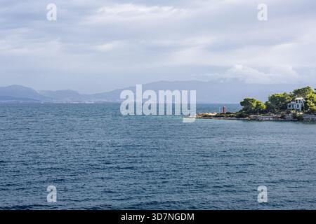 Eine ruhige Küstenszene mit einer felsigen Insel mit einem roten Leuchtturm, einem weißen Haus und grünen Bäumen am Ufer. Sanfte Wellen, ferne Berge, Stockfoto