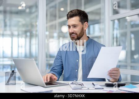 Junger bärtiger Unternehmer am Schreibtisch, lächelnd, während er an einem Laptop arbeitet und Geschäftsdokumente liest, was Produktivität, Selbstvertrauen und modernen beruflichen Erfolg vermittelt Stockfoto
