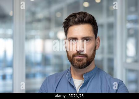 Selbstbewusster Mann mit Bart und ernstem Ausdruck, der in die Kamera blickt, steht in einem verschwommenen modernen Bürogebäude und steht für Professionalität und Geschäftserfolg Stockfoto
