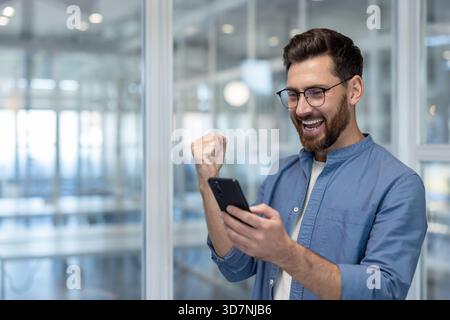 Junger Mann in blauem Hemd, der in einem modernen Büro steht und Freude und Begeisterung ausdrückt, während er auf sein Smartphone blickt und eine bedeutende Leistung oder gute Nachricht feiert Stockfoto