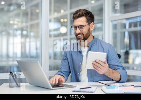 Junger Mann in Brille und lässigem Hemd lächelnd, Multitasking mit einem Laptop und einem digitalen Tablet, sitzt an einem weißen Schreibtisch in einem modernen, hell beleuchteten Büro und beschäftigt sich mit Technik Stockfoto
