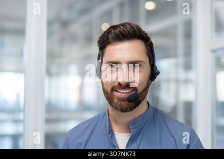 Junger bärtiger Mann mit einem selbstbewussten Lächeln, der ein Headset trägt und professionelle Kundenbetreuung und Kommunikationsdienste von einem modernen Callcenter-Büro aus bietet Stockfoto