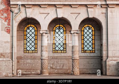 Drei Bogenfenster mit Buntglas, gestützt von kunstvollen Säulen, in einem verwitterten Steingebäude mit freiliegendem Mauerwerk. Stockfoto