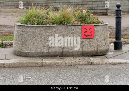 Straßenblumentopf aus Beton mit ornamentalem Gras und Rosen, mit einem roten Graffiti-Smiley-Schild an der Seite. Stockfoto