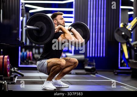 In einem modernen Fitnessstudio führt ein Mann eine Hocke mit einer Langhantel auf. Die lebendige Beleuchtung und die Ausstattung schaffen eine energiegeladene Atmosphäre für Krafttraining Stockfoto