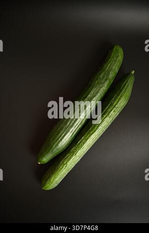 Top-down-Aufnahme von zwei langen, frischen grünen Gurken, die diagonal auf einem minimalistischen schwarzen Hintergrund platziert werden. Ideal für Themen der gesunden Ernährung, Veganismus und Stockfoto