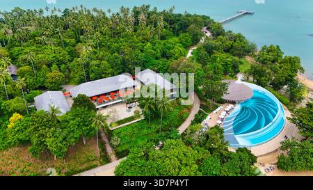 Blick aus der Vogelperspektive auf ein luxuriöses Resort inmitten üppiger tropischer Vegetation, mit einem markanten runden Pool und einem langen Pier, der sich in das türkisfarbene Meer erstreckt Stockfoto