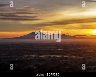 Aus der Vogelperspektive auf eine entfernte lawu-Bergkette, die sich vor einem lebhaften Sonnenaufgang mit Orange- und Gelbtönen im Kontrast zur dunklen Landschaft Be befindet Stockfoto