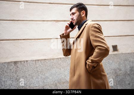 Ein gut gekleideter Mann in einem braunen Mantel geht auf einem glatten Gehweg entlang, in sein Telefongespräch vertieft. Er verströmt Selbstvertrauen, während er einen Mod hinuntergeht Stockfoto