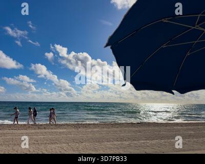 Strandbesucher spazieren an einem sonnigen Tag in Lauderdale-by-the-Sea unter einem hellen Morgenhimmel entlang der Küste, umrahmt vom Schatten eines großen Sonnenschirms Stockfoto