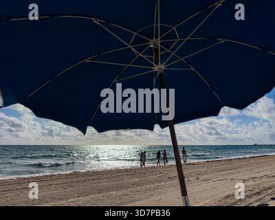 Strandbesucher spazieren an einem sonnigen Tag in Lauderdale-by-the-Sea unter einem hellen Morgenhimmel entlang der Küste, umrahmt vom Schatten eines großen Sonnenschirms Stockfoto