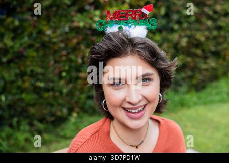 Frau lächelt, während sie im Hinterhof sitzt, trägt einen rostfarbenen Pullover und ein Haarreif von â€˜MERRY CHRISTMAS Stockfoto