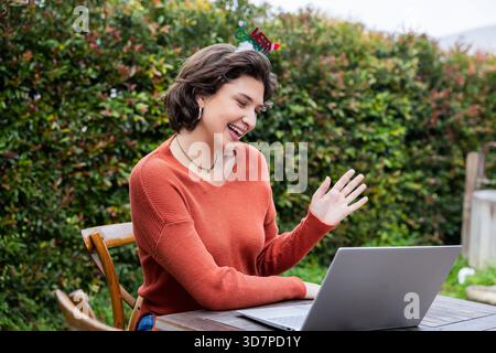 Frau, die am Laptop winkt und auf Holzstuhl am Holztisch sitzt, mit festlichem Stirnband im Hinterhof Stockfoto