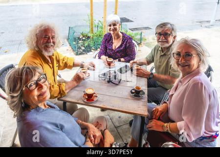 Lächelnde Gruppe von älteren Freunden versammelte sich an einem Café-Tisch im Freien und genoss Kaffee, Konversation und entspannte Zweisamkeit an einem sonnigen Tag in der Stadt, Div Stockfoto