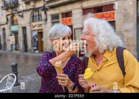 Seniorenpaar auf einer bezaubernden europäischen Straße, die Eis teilt, Frau füttert dem Mann einen Kegel, während sie schlendern, lächeln und einen süßen, romantischen Moment genießen Stockfoto