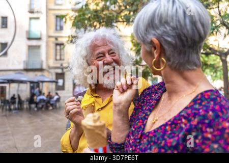 Seniorenpaar lacht und teilt Eis bei einem sonnigen Sommerdate auf einem belebten europäischen Stadtplatz, genießt lässigen Outdoor-Spaß, Zweisamkeit und Re Stockfoto