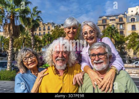 Glückliche, multirassische Seniorenfreunde, die während des Urlaubs auf einer sonnigen Stadtstraße lächeln und umarmen, die sich in den Ruhestand, Freundschaft und Aktivität freuen Stockfoto