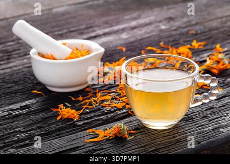 Eine Tasse Calendula-Tee sitzt auf einem rustikalen Holztisch, begleitet von getrockneten Blütenblättern und einem weißen Mörtel mit Blumen. Stockfoto