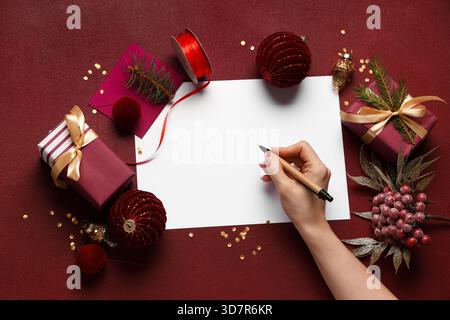 Weibliche Hand mit Weihnachtsdekorationen und Geschenkboxen, die Grußkarte auf burgunderrotem Hintergrund schreiben Stockfoto