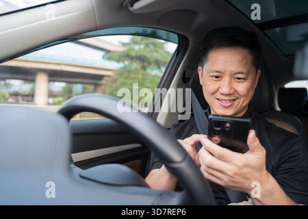 Ein Mann, der auf dem Fahrersitz sitzt, verwendet sein Smartphone, während das Fahrzeug angehalten wird, und hebt abgelenktes Fahrverhalten, den modernen Fahrzeuginnenraum und die hervor Stockfoto