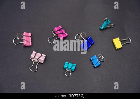 Eine Nahaufnahme von drei farbigen Binderclips, die aufrecht auf einer strukturierten, dunkelgrauen Oberfläche stehen. Stockfoto