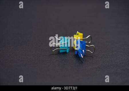 Eine Nahansicht von drei farbigen Binderclips (Petrol, Gelb und Blau), die aufrecht auf einer strukturierten, dunkelgrauen Oberfläche stehen. Stockfoto