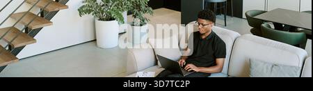 Junger Erwachsener Schwarzer Mann, der auf Sofa sitzt, mit Laptop im hellen Morgenlicht Stockfoto