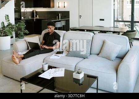 Junger Erwachsener Schwarzer Mann, der auf Sofa sitzt, mit Laptop im hellen Morgeninneren Stockfoto