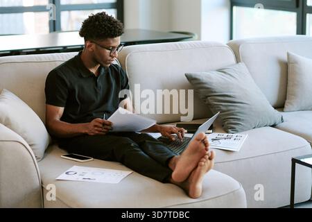 Junger Erwachsener Schwarzer Mann, der am Morgen zu Hause ferngesteuert am Laptop arbeitet Stockfoto