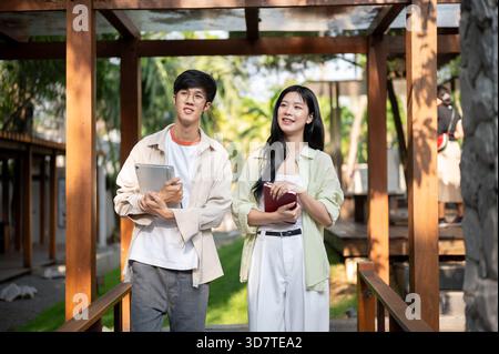 Lächelnde asiatische Freunde oder ein Mann und eine Frau, die Bücher halten und zusammen im Holzkorridor stehen. Arbeiten oder Lernen im Freien, Garden Cafe oder Restaurant. Stockfoto