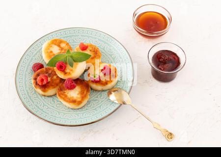Hausgemachte Quark-Krapfen mit Himbeeren und einem Teelöffel. Glasschalen mit Erdbeermarmelade und Honig auf weißem Hintergrund. Draufsicht. Stockfoto