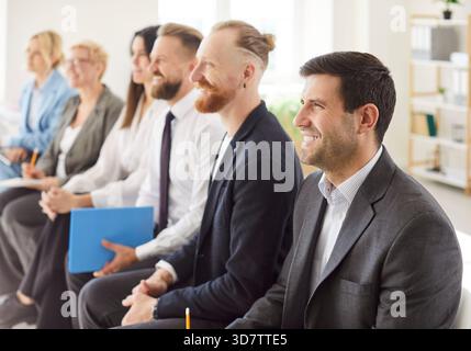 Eine Gruppe fröhlich lächelnder Geschäftsleute sitzt in einer Reihe auf Stühlen und hört ihrem Kollegen zu Stockfoto