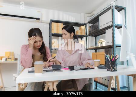 Zwei junge Frauen arbeiten in einem kleinen Büro zusammen, organisieren Pakete, prüfen Finanzberichte und verwalten Bestellungen auf digitalen Geräten Stockfoto