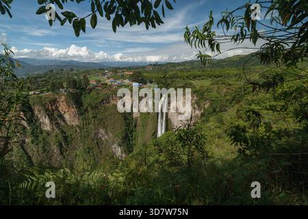 Malerischer Blick auf einen Wasserfall, der in einen üppig grünen Canyon mit einem kleinen Dorf in der Nähe fällt. San Agustin, Kolumbien Stockfoto
