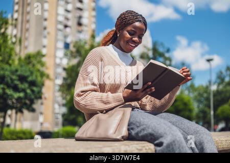Fröhliche Afroamerikanerin, die sich im Stadtpark entspannt und ein Buch liest. Stockfoto