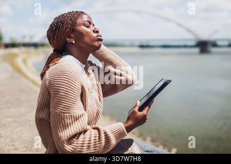 Die junge besorgte schwarze Frau benutzt ein digitales Tablet. Sie sitzt am Fluss in der Stadt. Stockfoto