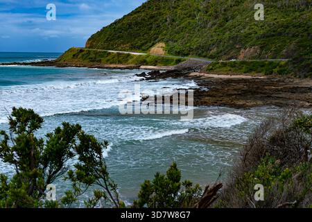 Blick auf die Wellen, die auf die Felsen am Strand stürzen, mit der Great Ocean Road und dem grünen Berg, Victoria, Australien. Stockfoto