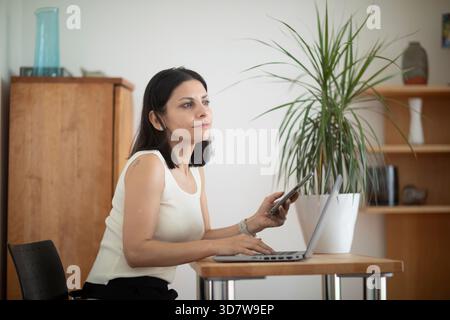 Eine Frau in einem weißen Oberteil sitzt an einem Schreibtisch mit einem Laptop und hält ein Telefon. Deutschland Stockfoto