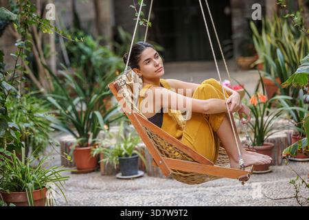 Frau in gelbem Outfit, die auf einer Schaukel sitzt, umgeben von üppigen grünen Pflanzen. Sierra Alhamilla, Almeria, Spanien Stockfoto