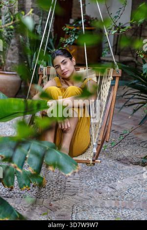 Frau in gelbem Kleid sitzt auf einem Hängesessel umgeben von grünen Pflanzen Sierra Alhamilla, Almeria, Spanien Stockfoto