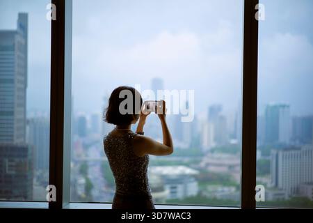 Frau, die an einem bewölkten Tag durch ein großes Fenster ein Foto von einer Skyline der Stadt macht. Shanghai, China Stockfoto