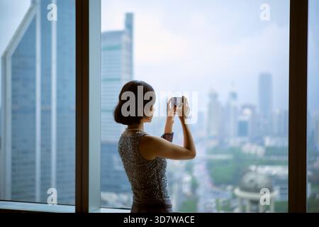 Frau, die ein Foto von einer Skyline der Stadt von einem hohen Aussichtspunkt in der Nähe eines Fensters macht. Shanghai, China Stockfoto