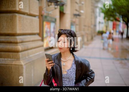 Eine junge Frau in einer Stadtstraße überprüft ihr Handy, trägt Sonnenbrille und modische Kleidung. Shanghai, China Stockfoto