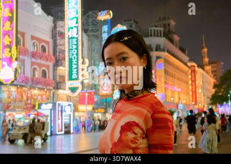Eine junge Frau in einem bunten Outfit, die nachts auf einer hellen, geschäftigen Stadtstraße steht. Shanghai, China Stockfoto