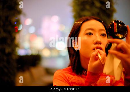Junge Frau, die Lippenstift in einer Stadtlandschaft mit verschwommenen bunten Lichtern in der Nacht aufträgt. Shanghai, China Stockfoto